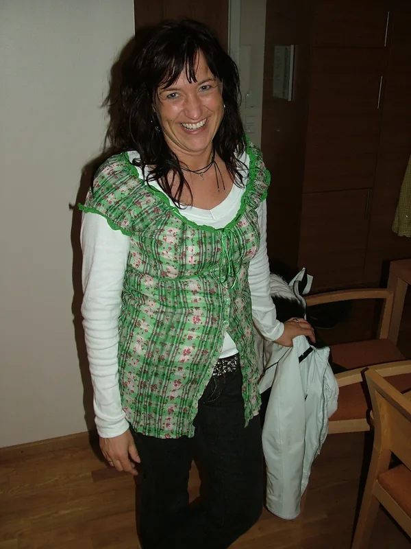Femme souriante aux cheveux bruns portant une veste verte à motifs, un haut blanc et un jean, tenant une veste blanche.