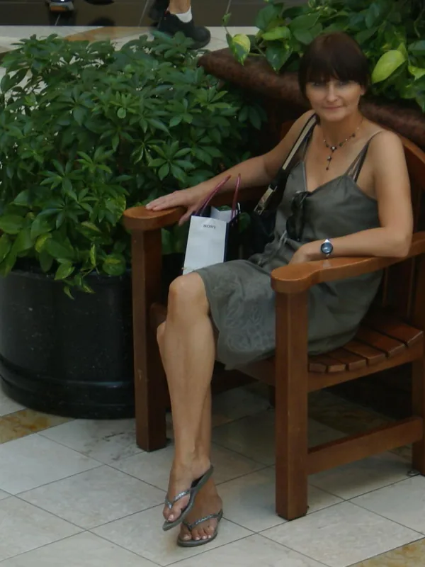 Femme assise sur une chaise en bois dans un hall, entourée de plantes vertes, portant une robe grise et des sandales.