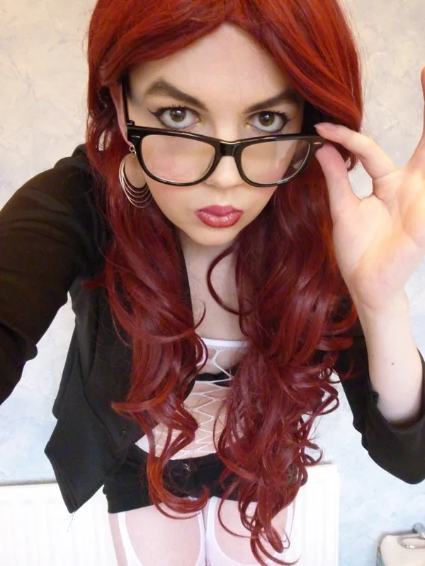 Portrait d'une femme aux cheveux roux portant des lunettes, une veste noire et un haut blanc, regardant l'objectif.