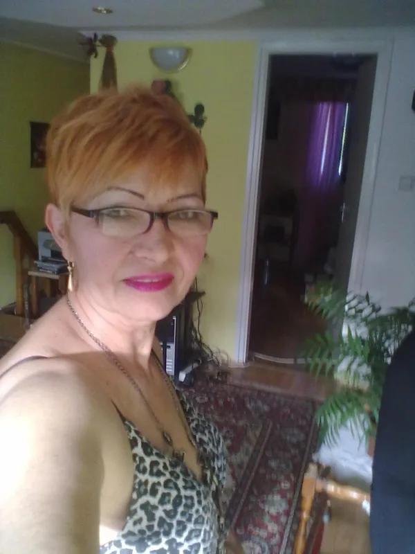 Portrait d'une femme aux cheveux courts roux, portant des lunettes et un haut léopard, dans un salon intérieur.
