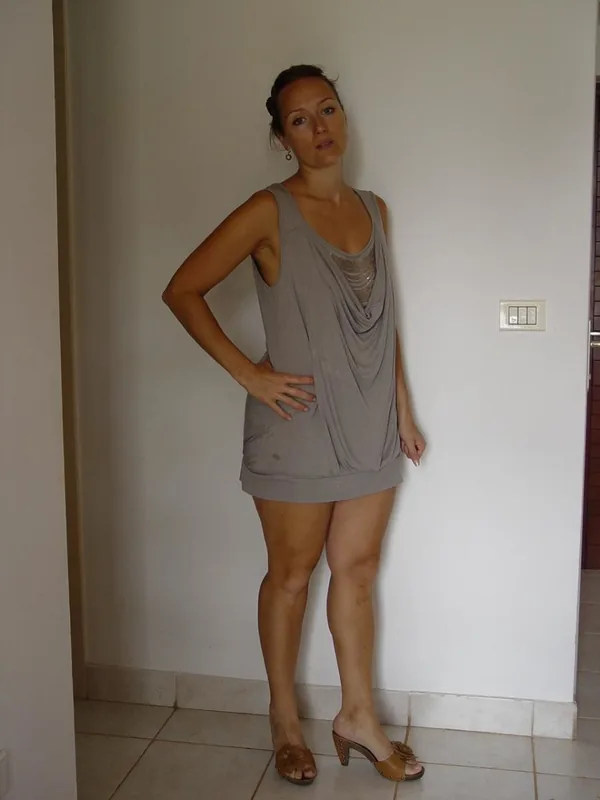 Femme debout en tenue décontractée, portant un haut gris et des sandales, posant contre un mur blanc.