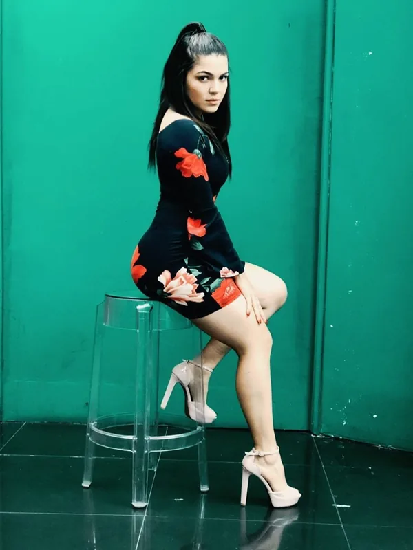 Femme assise sur un tabouret transparent devant un mur vert, portant une robe noire à fleurs et des talons hauts.