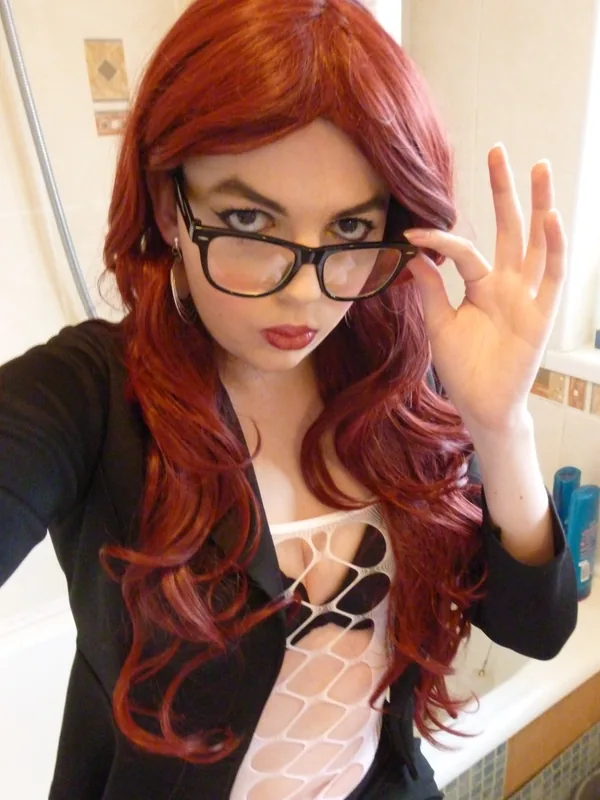 Portrait d'une femme aux cheveux roux portant des lunettes noires, une veste noire et un haut blanc transparent, dans une salle de bain.