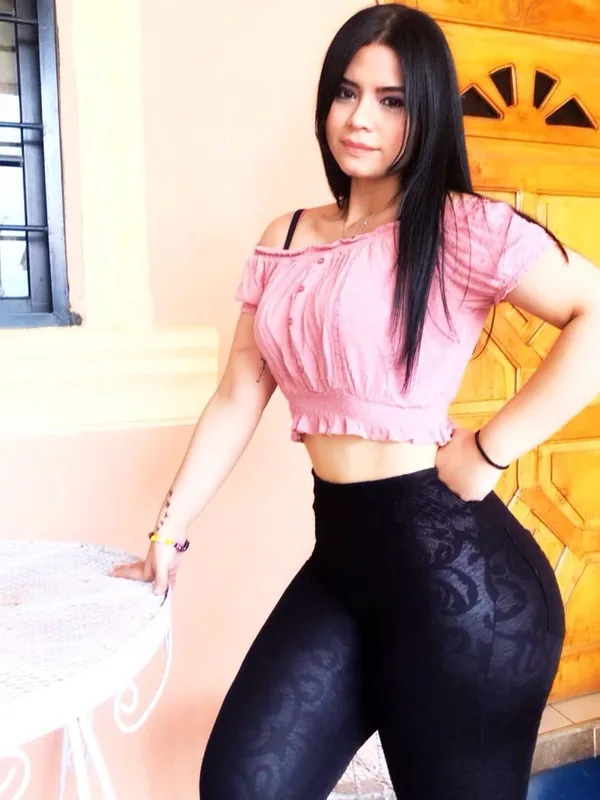 Femme aux cheveux noirs portant un haut rose et des leggings noirs, posant près d'une table blanche devant une porte en bois.