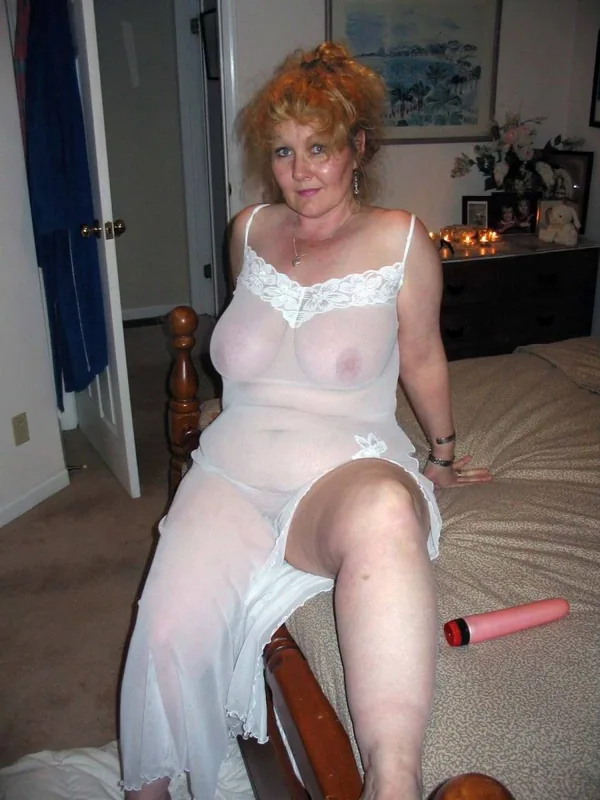 Femme aux cheveux roux assise sur un lit, vêtue d'une chemise de nuit transparente blanche, avec un jouet rose à côté d'elle.