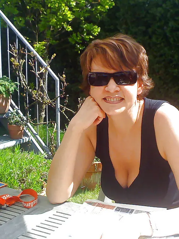 Femme portant des lunettes de soleil et un haut noir, assise à une table en extérieur dans un jardin ensoleillé.