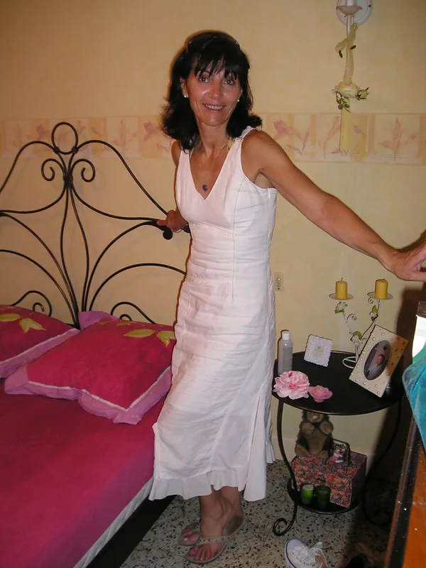 Femme souriante en robe blanche debout près d'un lit rose dans une chambre décorée.