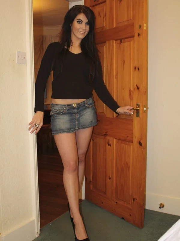Femme aux cheveux bruns portant un haut noir et une jupe en jean, debout dans un encadrement de porte en bois.