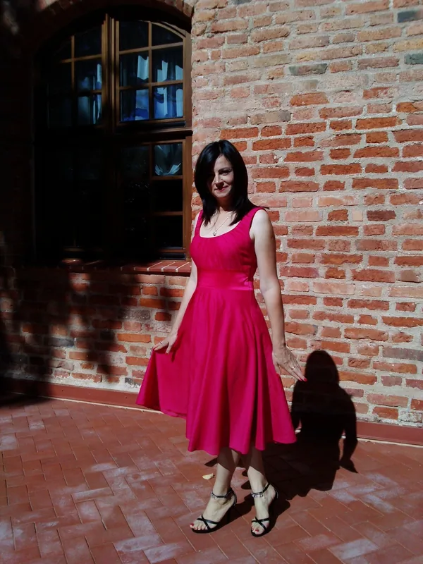 Femme en robe rose vif posant devant un mur de briques sous la lumière du soleil.