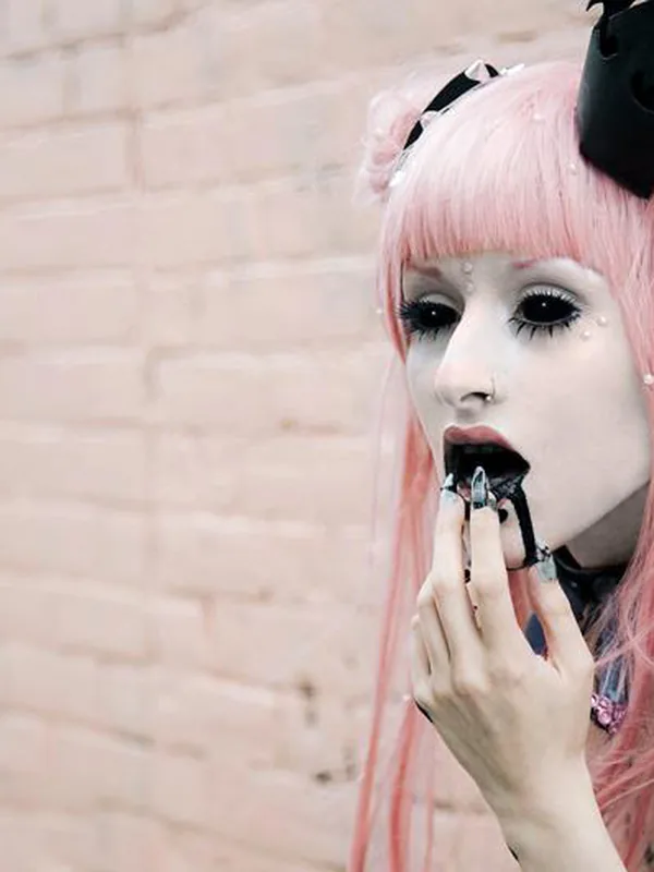 Portrait d'une personne aux cheveux roses, maquillage blanc, oreilles pointues et couronne noire, devant un mur rose.